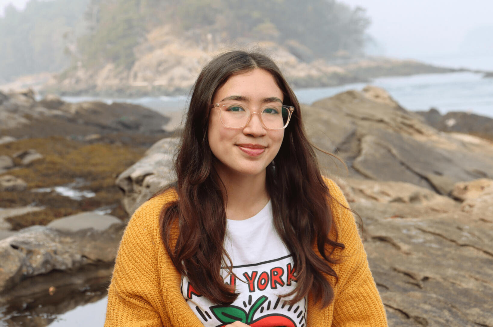 Sofia Osborne sitting on rocks by the ocean wearing a yellow sweater and glasses.